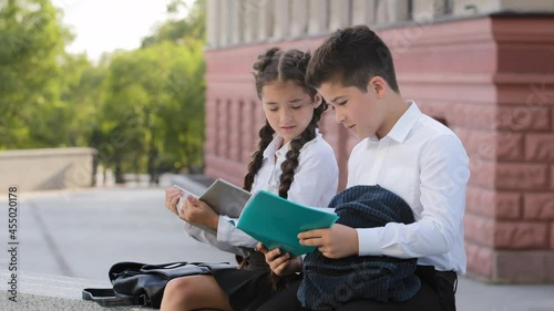 Two hispnic children students pupils sitting outdoors with books reading after class. Girl reads book sad boy showing low bad mark in diary mistake notebook talk about problem failure, back to school photo