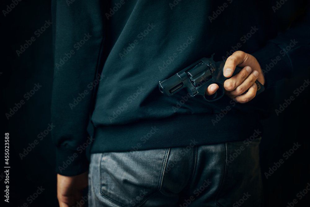 Criminal person with revolver on his back. Man in dark clothing is holding gun. Firearm for defense or attack. Murderer in black background.