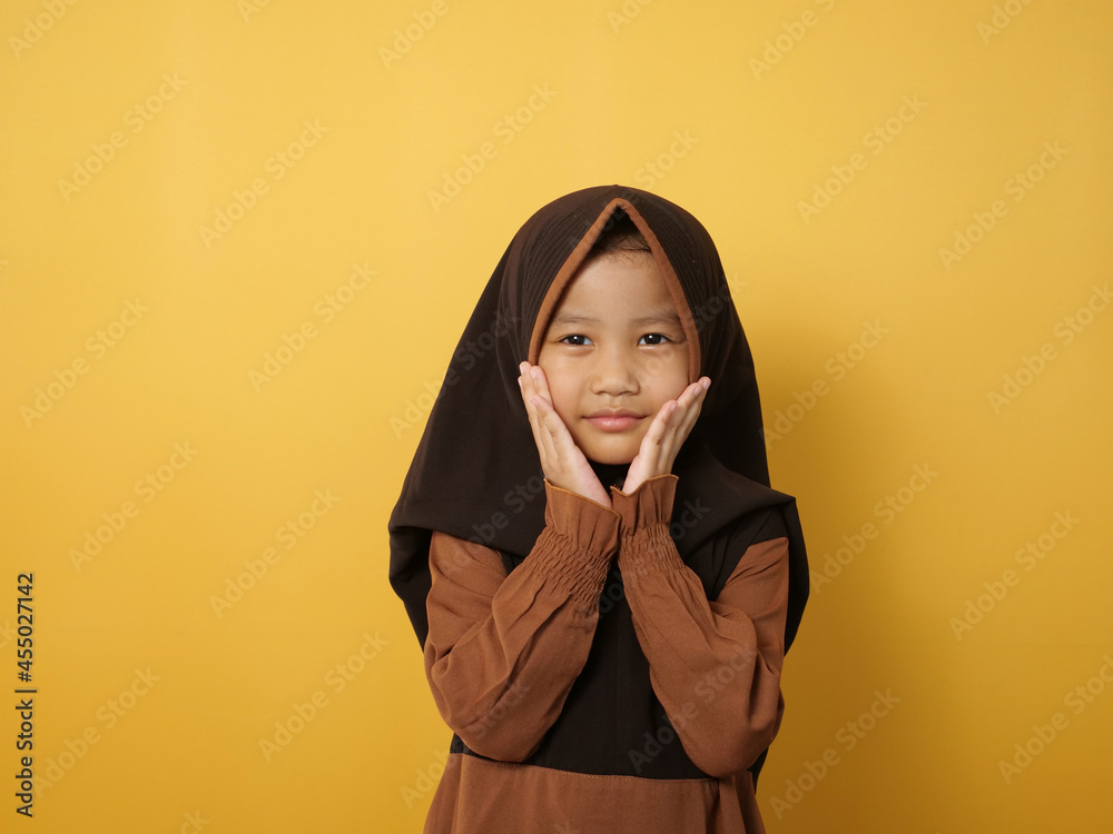Funny cute Asian muslim little girl wearing hijab looking at camera and smiling, against yellow Funny cute Asian muslim little girl wearing hijab looking at camera and smiling, against yellow