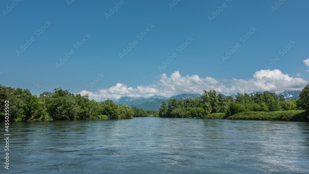 A peaceful blue river flows between the banks with lush green vegetation. In the distance, against the background of the azure sky and clouds, a mountain range is visible. Kamchatka A peaceful blue river flows between the banks with lush green vegetation. In the distance, against the background of the azure sky and clouds, a mountain range is visible. Kamchatka