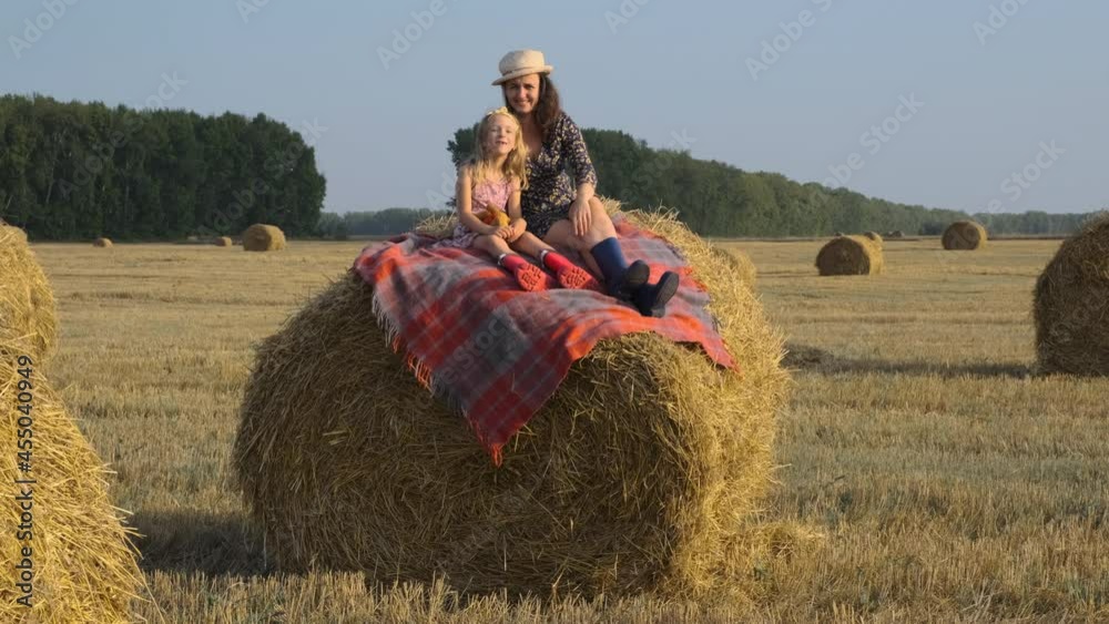 Young Mother with Little Daughter Sitting on a Hay Bale in a Harvested Field. Happy Childhood and Freedom Concept Young Mother with Little Daughter Sitting on a Hay Bale in a Harvested Field. Happy Childhood and Freedom Concept