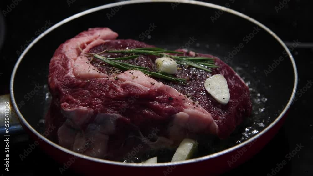 Close-up view of chuck steak frying on frying pan with garlic and rosemary branches. Beef meat cooking. Selective focus. 4K resolution video. Food preparation theme. Close-up view of chuck steak frying on frying pan with garlic and rosemary branches. Beef meat cooking. Selective focus. 4K resolution video. Food preparation theme.