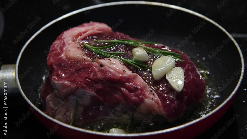 Close-up view of chuck steak frying on frying pan with garlic and rosemary branches. Beef meat cooking. Selective focus. 4K resolution video. Food preparation theme. Close-up view of chuck steak frying on frying pan with garlic and rosemary branches. Beef meat cooking. Selective focus. 4K resolution video. Food preparation theme.