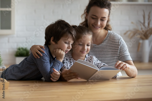 Happy young mom and two sibling kids involved in fairy tale, reading paper book together at kitchen table. Mother telling fantasy story to kids, improving interest to literature, helping with homework