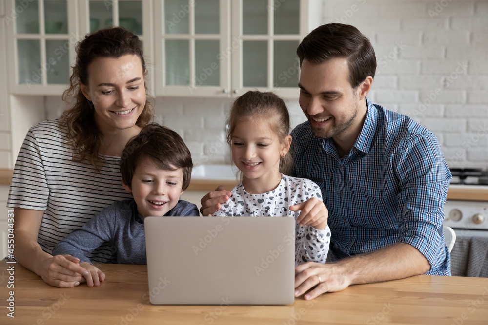 Happy joyful family using laptop in kitchen, enjoying leisure time together, watching funny movie online. Couple of parents and two kids making video call, shopping, booking hotel on internet