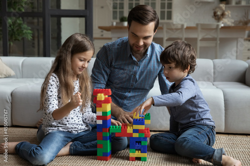 Happy best dad and two sibling children playing together on warm floor  building toy tower on clean carpet  constructing model from plastic blocks  improving creativity in game. Family home activity