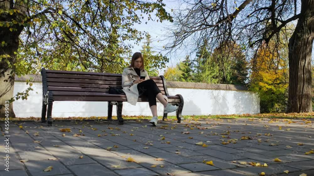 A young woman sits on a bench in an autumn park and sends a message on the phone.