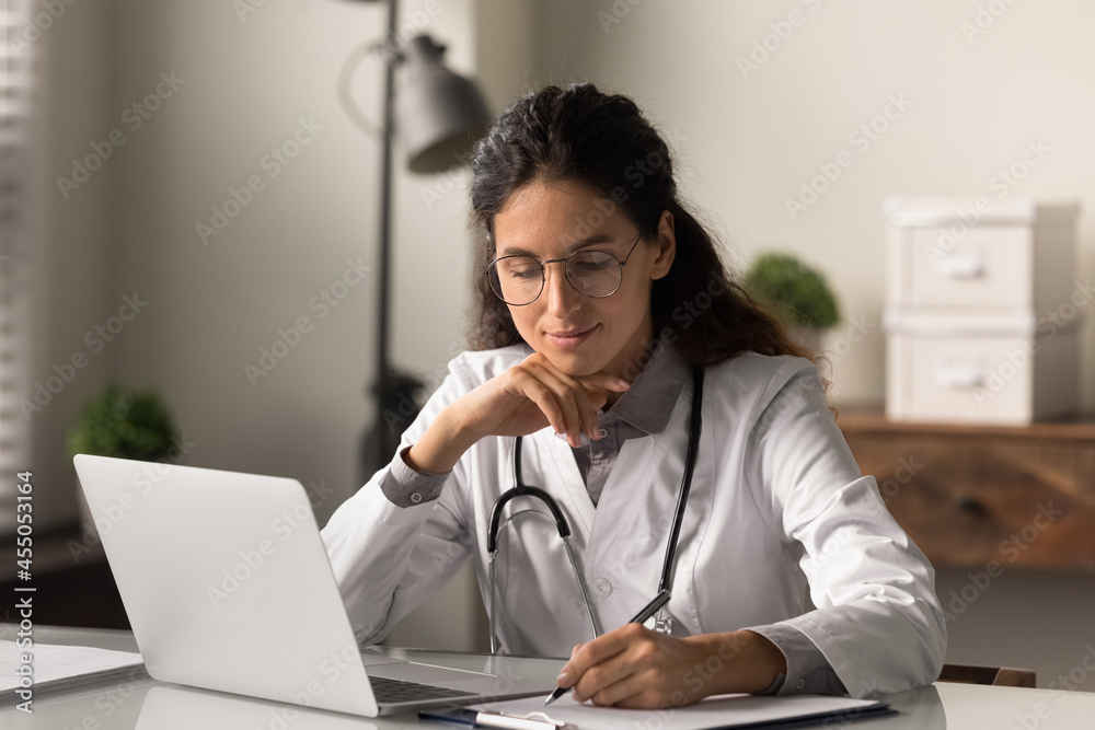 Focused smiling GP doctor giving online distance consultation on laptop, listening to patient complaints, taking notes during video call. Physician writing reports at workplace with computer Focused smiling GP doctor giving online distance consultation on laptop, listening to patient complaints, taking notes during video call. Physician writing reports at workplace with computer