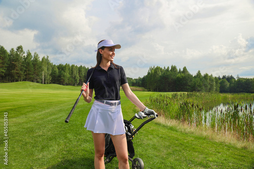 Woman in stylish summer golf outfit walking with bag of drivers on beautiful green course for golf game.