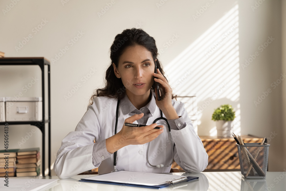 Serious female doctor giving telephone consultation, talking to patient on cellphone, making phone call from clinic office. General practitioner working on healthcare hotline, answering questions Serious female doctor giving telephone consultation, talking to patient on cellphone, making phone call from clinic office. General practitioner working on healthcare hotline, answering questions