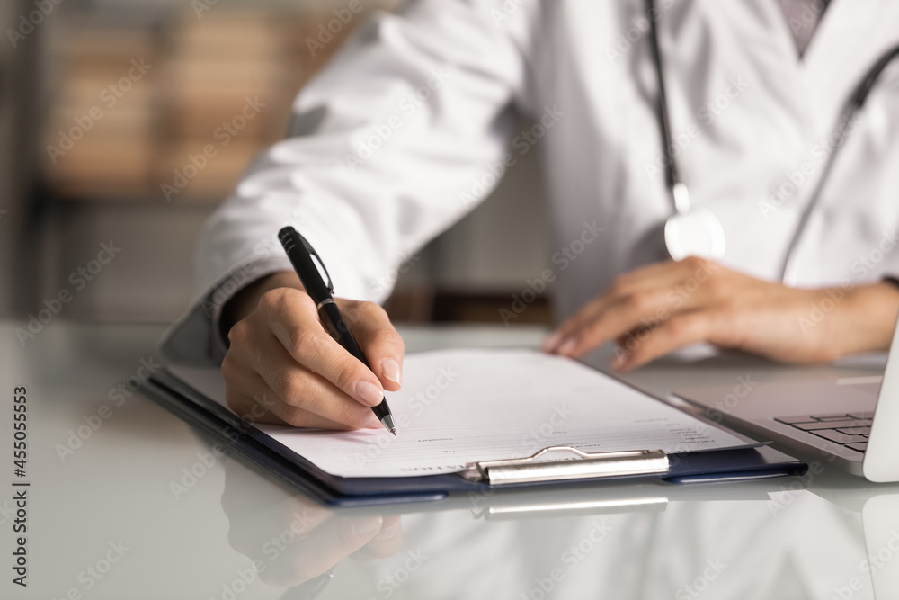 Hands of general practitioner filling paper medical records. Doctor in white coat doing paperwork at workplace with laptop, writing notes, preparing documents, reports, prescription. Close up