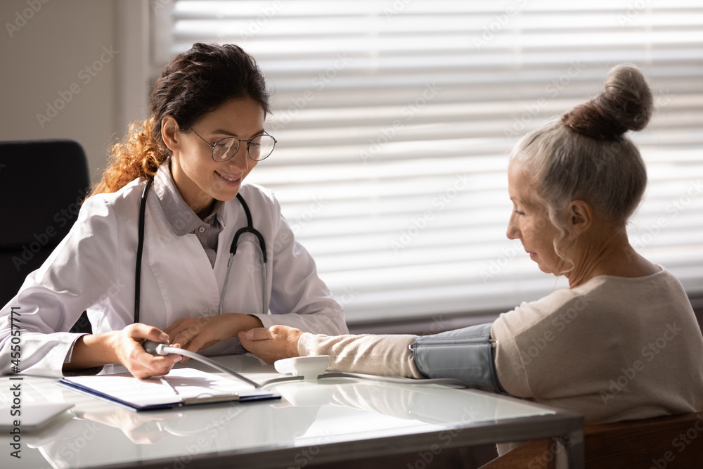 Smiling friendly doctor checking blood pressure of older female patient at appointment in office, using sphygmomanometer, examining woman with hypertension or hypotension, fast heartbeat Smiling friendly doctor checking blood pressure of older female patient at appointment in office, using sphygmomanometer, examining woman with hypertension or hypotension, fast heartbeat