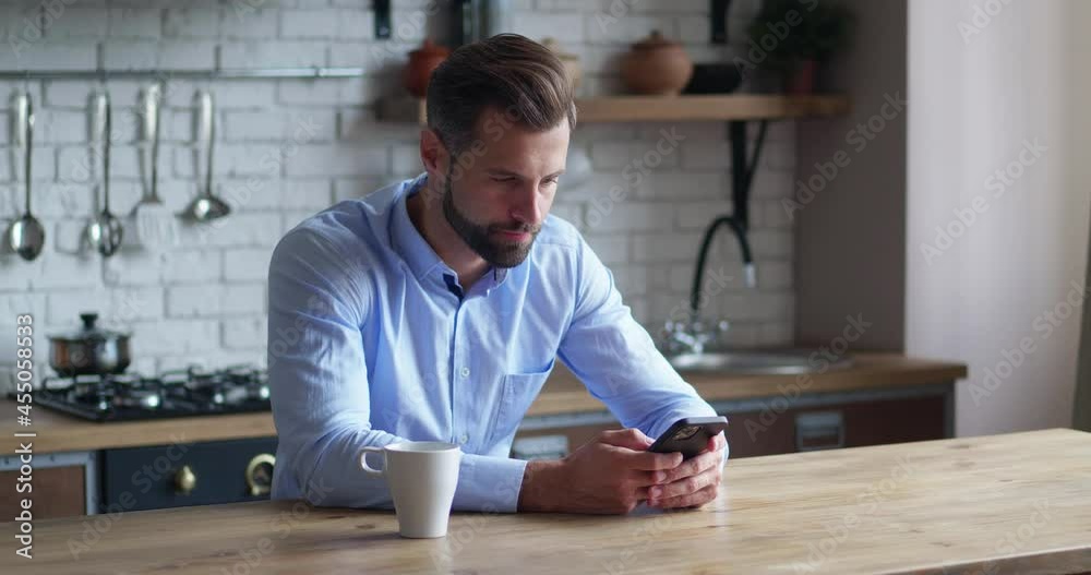 Bearded male millennial professional holding modern smartphone texting message in kitchen. Young businessman using helpful mobile apps for business time management organization concept sitting at work