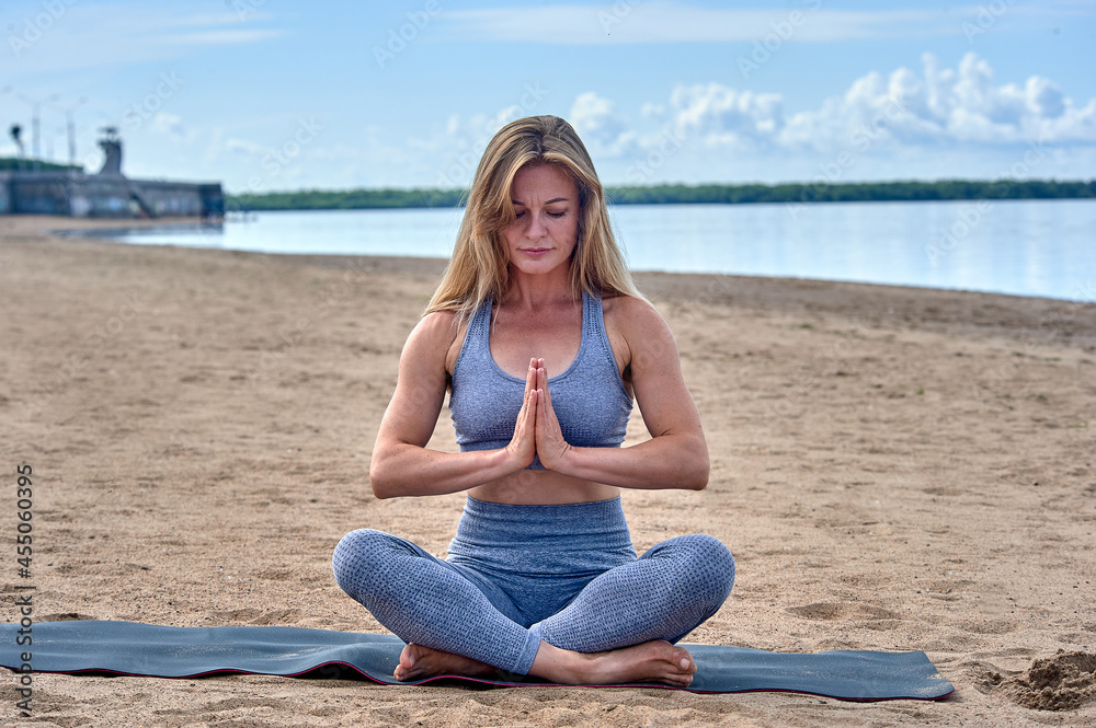 A woman sits in a lotus position on a sandy beach against the background of a river, yoga