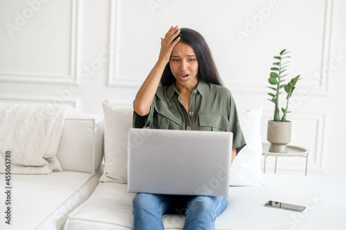 Upset and frustrated young indian woman using laptop for remote work from home, nervous about mistakes, stressful job, mixed-race female student has problem with task holding head with desperation photo