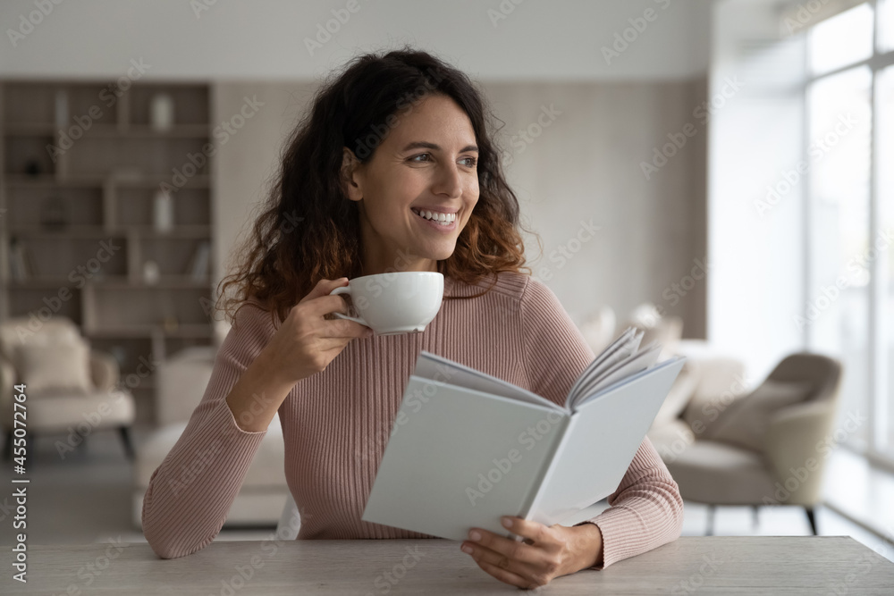 Happy beautiful young hispanic woman drinking cup of hot tea or coffee, reading favorite novel literature in paper book or bestseller, enjoying peaceful domestic weekend pastime activity at home. Happy beautiful young hispanic woman drinking cup of hot tea or coffee, reading favorite novel literature in paper book or bestseller, enjoying peaceful domestic weekend pastime activity at home.