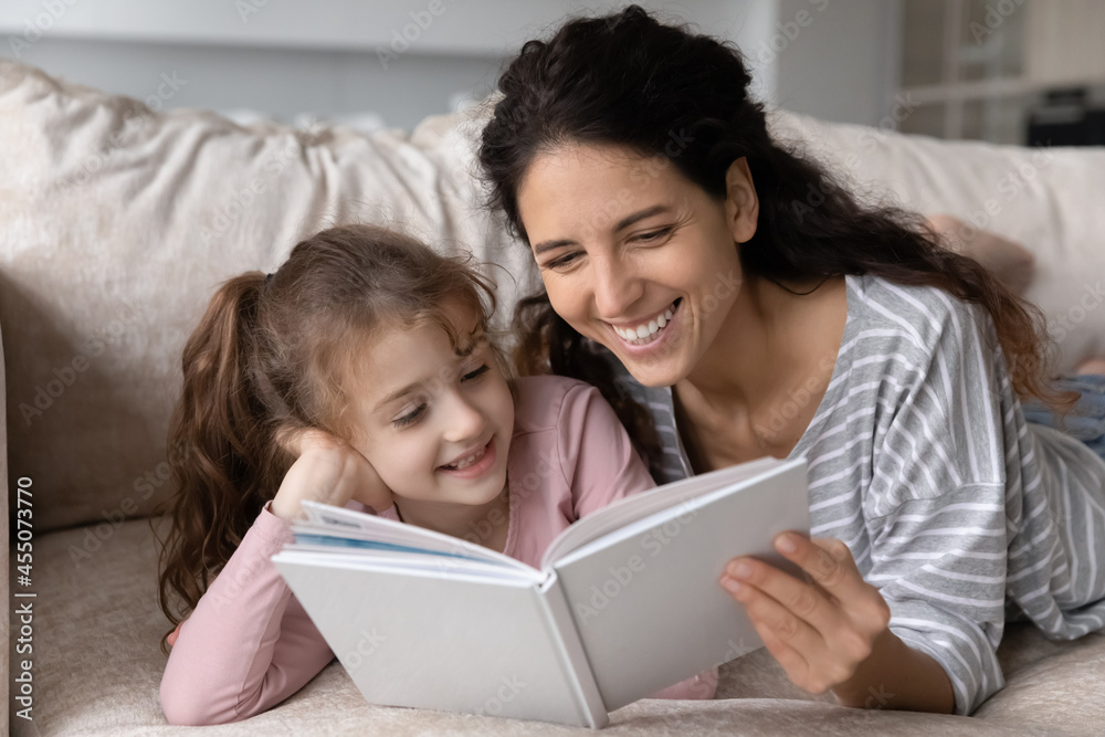 Happy bonding two female generations family lying on comfortable sofa, reading funny stories in paper book. Smiling young mother enjoying leisure activity together with cute child daughter at home. Happy bonding two female generations family lying on comfortable sofa, reading funny stories in paper book. Smiling young mother enjoying leisure activity together with cute child daughter at home.