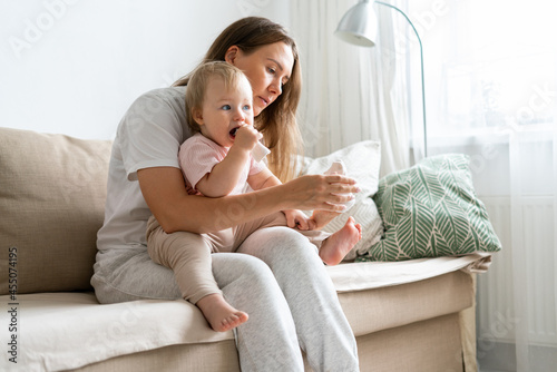 Side view of caucasian babysitter in casual clothes sitting on sofa with curious blond blue-eyed child and putting socks on baby feet at home. Concept of child care and parenthood. Copy space