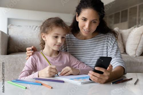 Smiling young mother showing images examples on cellphone screen or watching educational video to adorable little child daughter, redrawing pictures together in paper album, hobby activity concept.