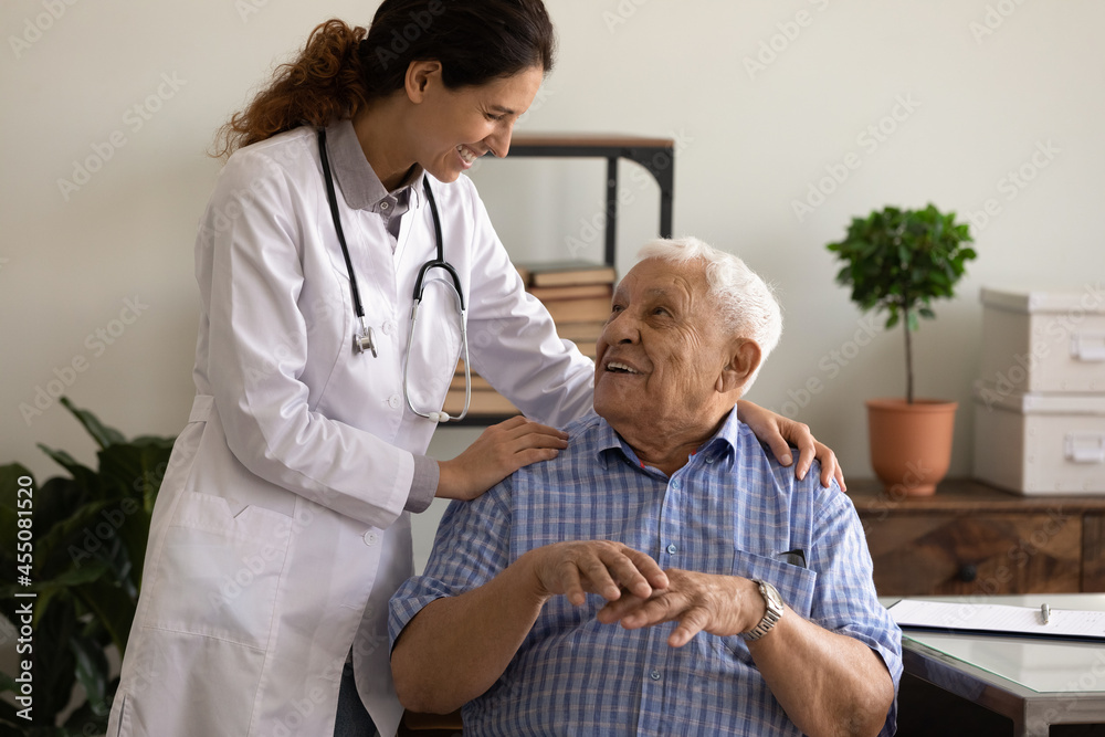 Obraz premium Happy young female GP doctor giving hug and support to optimistic old 80s patient. Senior man visiting, consulting therapist, discussing successful treatment, recovery, remission, good health