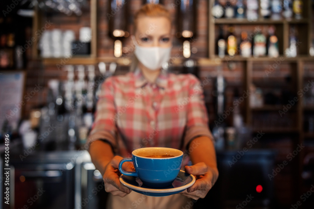 The waitress offered a cup of coffee. A photo of an isolated blue cup of coffee behind a blurred background of a woman with a protective face mask. Female hands hold the cup and extend it to the guest The waitress offered a cup of coffee. A photo of an isolated blue cup of coffee behind a blurred background of a woman with a protective face mask. Female hands hold the cup and extend it to the guest