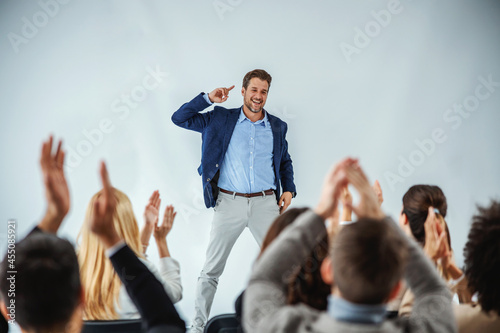 Smiling motivational speaker standing in front of his audience who is clapping.