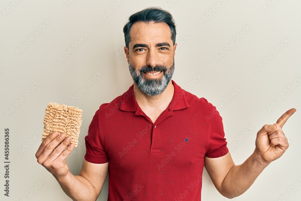 Middle age man with beard and grey hair holding uncooked noodles smiling happy pointing with hand and finger to the side Middle age man with beard and grey hair holding uncooked noodles smiling happy pointing with hand and finger to the side