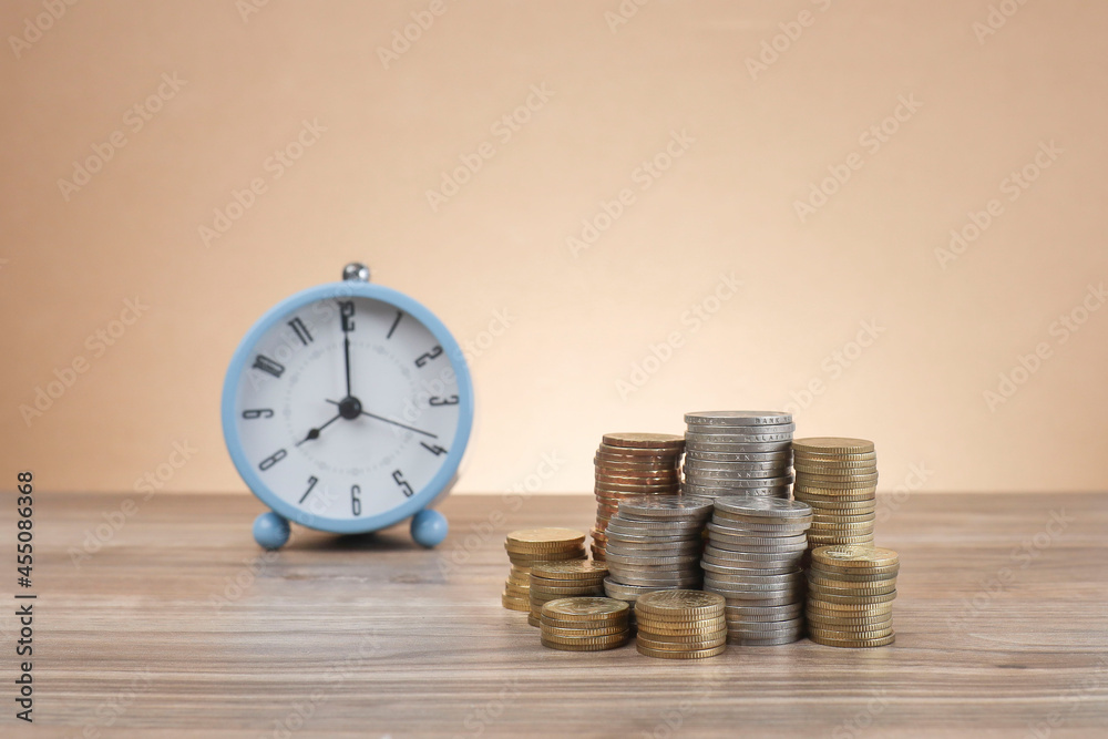 Business and time concept. Coins stacked on the wooden table with an alarm clock at the back Business and time concept. Coins stacked on the wooden table with an alarm clock at the back