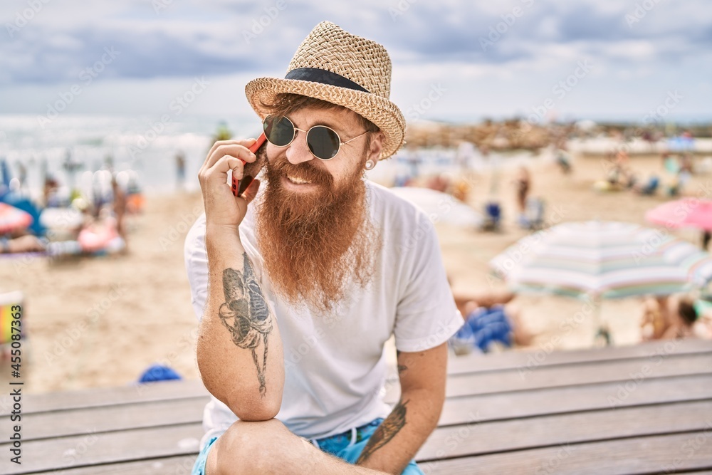 Young redhead tourist man talking on the smartphone sitting on the bench at the beach.