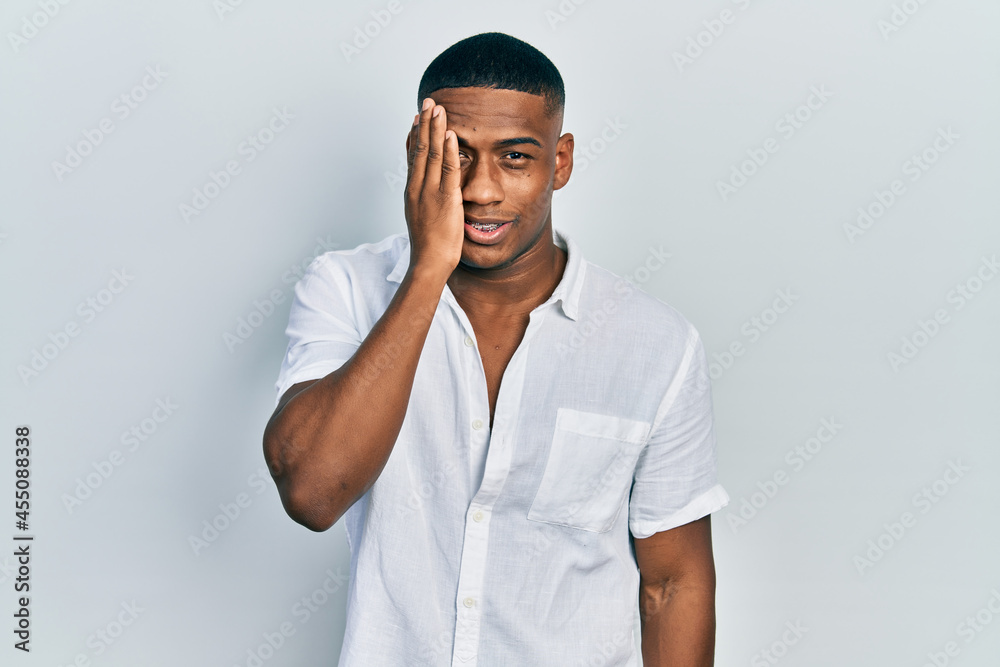 Young black man wearing casual white shirt covering one eye with hand, confident smile on face and surprise emotion. Young black man wearing casual white shirt covering one eye with hand, confident smile on face and surprise emotion.