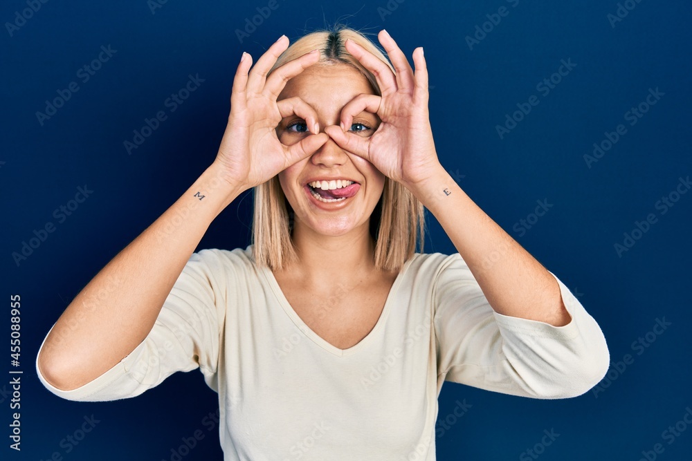 Beautiful blonde woman wearing casual sweater doing ok gesture like binoculars sticking tongue out, eyes looking through fingers. crazy expression. Beautiful blonde woman wearing casual sweater doing ok gesture like binoculars sticking tongue out, eyes looking through fingers. crazy expression.