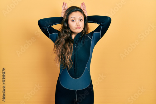 Young hispanic girl wearing diver neoprene uniform doing bunny ears gesture with hands palms looking cynical and skeptical. easter rabbit concept.