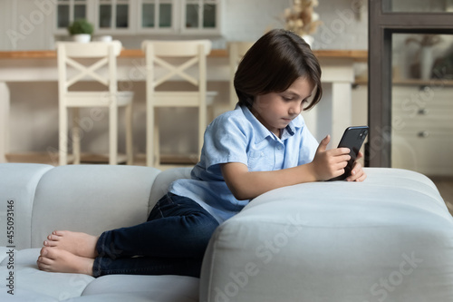Full length relaxed happy little kid boy using cellphone apps, sitting alone on cozy sofa, enjoying playing games, watching funny videos, spending time online in social networks, modern tech addiction
