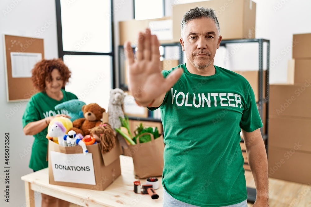 Middle age man wearing volunteer t shirt at donations stand doing stop sing with palm of the hand. warning expression with negative and serious gesture on the face. Middle age man wearing volunteer t shirt at donations stand doing stop sing with palm of the hand. warning expression with negative and serious gesture on the face.