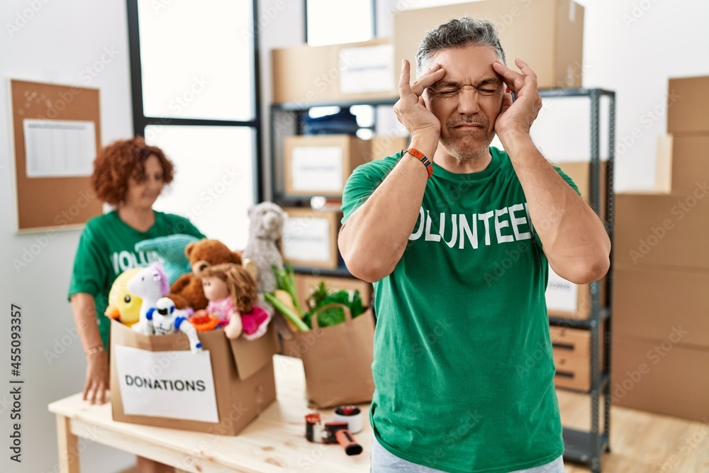 Middle age man wearing volunteer t shirt at donations stand with hand on head for pain in head because stress. suffering migraine. Middle age man wearing volunteer t shirt at donations stand with hand on head for pain in head because stress. suffering migraine.