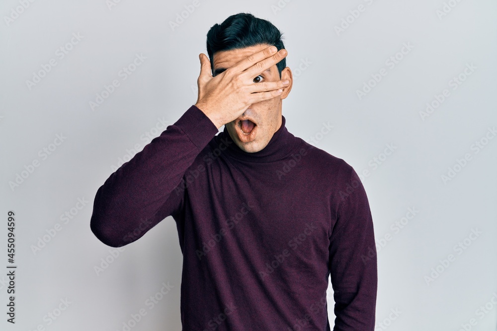 Handsome hispanic man wearing turtleneck sweater peeking in shock covering face and eyes with hand, looking through fingers afraid