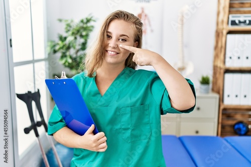 Young caucasian woman working at pain recovery clinic pointing with hand finger to face and nose  smiling cheerful. beauty concept