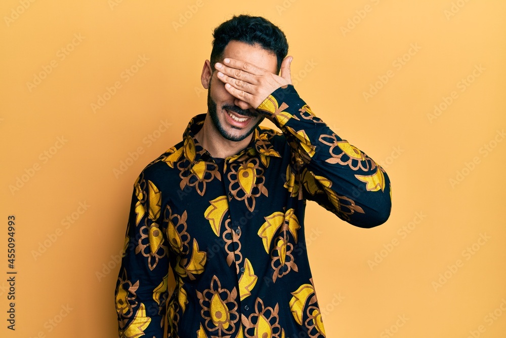 Young hispanic man wearing casual shirt smiling and laughing with hand on face covering eyes for surprise. blind concept. Young hispanic man wearing casual shirt smiling and laughing with hand on face covering eyes for surprise. blind concept.