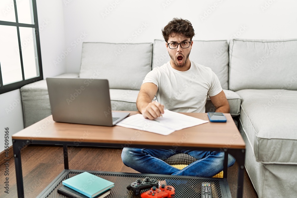 Hispanic man doing papers at home in shock face, looking skeptical and sarcastic, surprised with open mouth Hispanic man doing papers at home in shock face, looking skeptical and sarcastic, surprised with open mouth