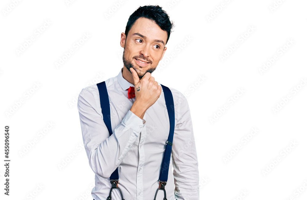 Hispanic man with beard wearing hipster look with bow tie and suspenders with hand on chin thinking about question, pensive expression. smiling with thoughtful face. doubt concept. Hispanic man with beard wearing hipster look with bow tie and suspenders with hand on chin thinking about question, pensive expression. smiling with thoughtful face. doubt concept.