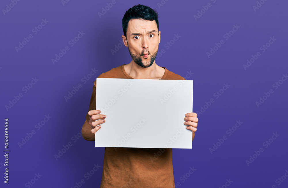 Hispanic man with beard holding blank empty banner making fish face with mouth and squinting eyes, crazy and comical. Hispanic man with beard holding blank empty banner making fish face with mouth and squinting eyes, crazy and comical.