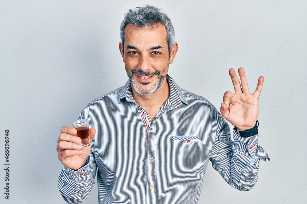 Handsome middle age man with grey hair drinking whiskey shot doing ok sign with fingers, smiling friendly gesturing excellent symbol Handsome middle age man with grey hair drinking whiskey shot doing ok sign with fingers, smiling friendly gesturing excellent symbol