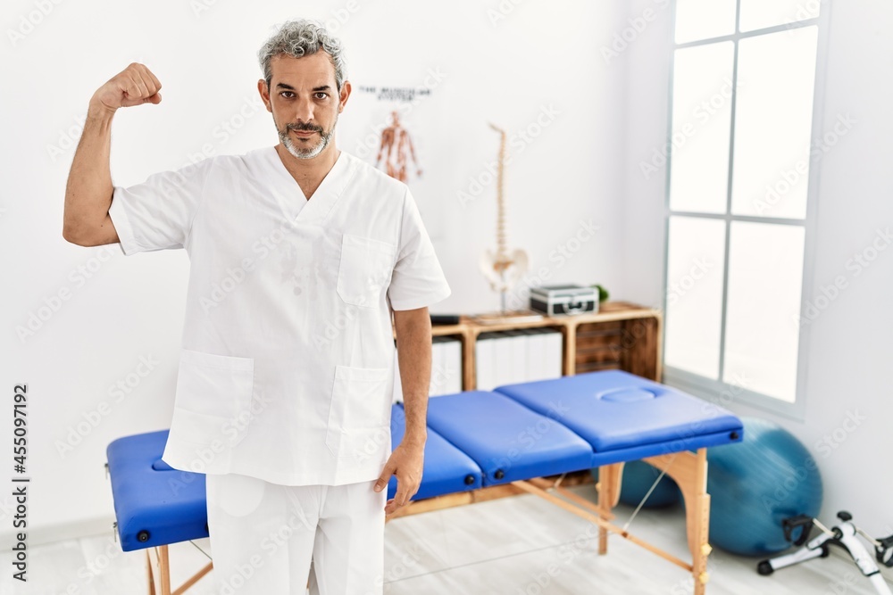 Middle age hispanic therapist man working at pain recovery clinic strong person showing arm muscle, confident and proud of power Middle age hispanic therapist man working at pain recovery clinic strong person showing arm muscle, confident and proud of power