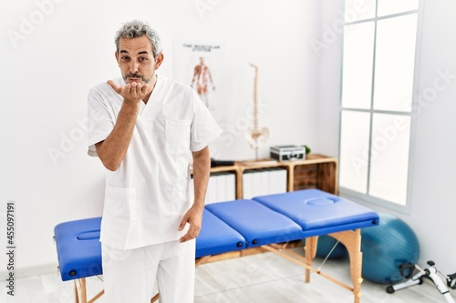 Middle age hispanic therapist man working at pain recovery clinic looking at the camera blowing a kiss with hand on air being lovely and sexy. love expression.