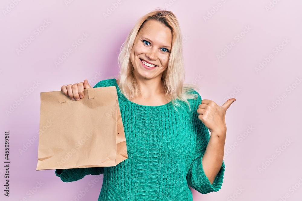 Beautiful caucasian blonde woman holding take away paper bag pointing thumb up to the side smiling happy with open mouth Beautiful caucasian blonde woman holding take away paper bag pointing thumb up to the side smiling happy with open mouth