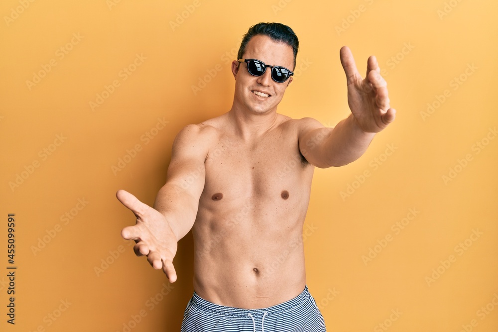 Handsome young man wearing swimsuit and sunglasses looking at the camera smiling with open arms for hug. cheerful expression embracing happiness.