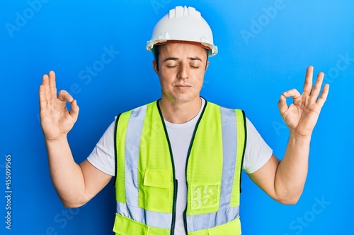 Handsome young man wearing safety helmet and reflective jacket relax and smiling with eyes closed doing meditation gesture with fingers. yoga concept.