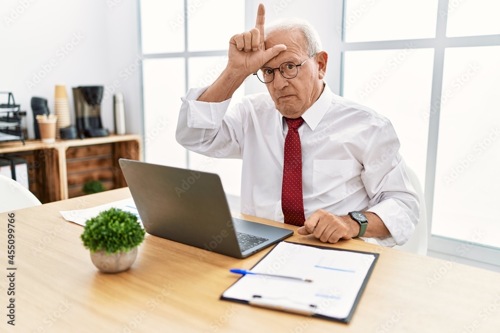 Senior man working at the office using computer laptop making fun of people with fingers on forehead doing loser gesture mocking and insulting. Senior man working at the office using computer laptop making fun of people with fingers on forehead doing loser gesture mocking and insulting.
