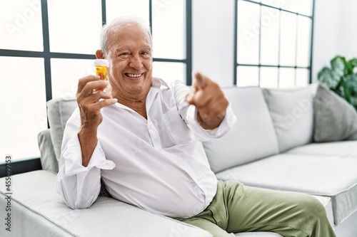 Senior man holding pills pointing to you and the camera with fingers, smiling positive and cheerful