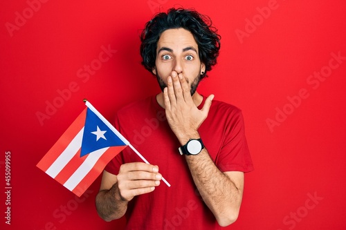 Handsome hispanic man holding puerto rico flag covering mouth with hand, shocked and afraid for mistake. surprised expression